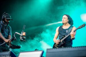 Guitarist with eyes closed plays a black electric guitar on a teal-lit stage while a camera operator with headset and rig stands to the left.