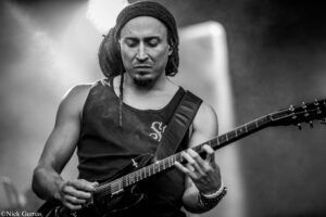 Male guitarist playing a black electric guitar on stage, eyes closed, wearing a head wrap and sleeveless shirt in black-and-white. There’s a watermark in the bottom-left corner.