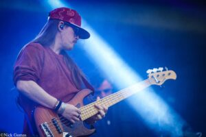 Guitarist playing a bass on stage under blue lights, wearing a red patterned cap and maroon sweater.