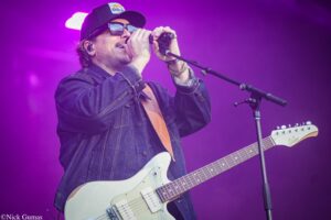 Male singer wearing a cap and sunglasses, singing into a microphone while playing a white electric guitar on a purple-lit stage.