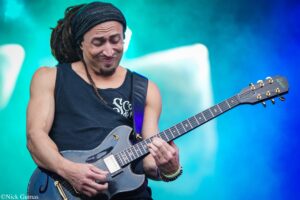 Male guitarist with dreadlocks in a black sleeveless shirt playing a gray electric guitar onstage, colorful teal-blue backdrop behind him.
