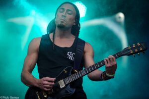 Guitarist performing on stage with teal lighting, wearing a black sleeveless shirt and dreadlocks, eyes closed as they play a black electric guitar.