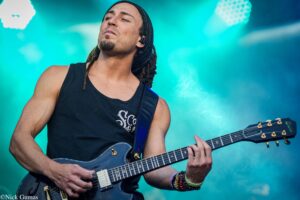 Male guitarist wearing a black sleeveless shirt, playing a dark electric guitar on a teal-lit stage.