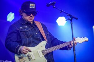 Guitarist wearing a denim jacket and cap with sunglasses performing on a blue-lit stage.