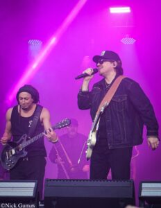 Two guitarists on a purple-lit stage, one singing into a microphone while the other plays a guitar.