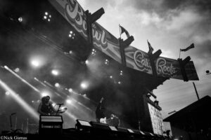 Black-and-white photo of a live outdoor concert with musicians on a smoky stage and bright lights overhead.