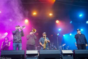 A live band on stage performing with brass and wind instruments under colorful purple, pink, and blue lights and stage fog.