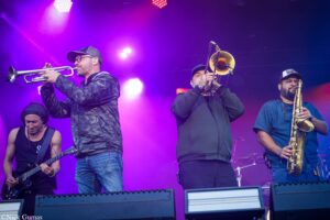 Four musicians performing on a stage: guitarist left, trumpeter and trombonist center, and saxophonist right, under vibrant purple stage lights.