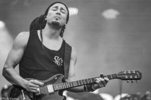 Male guitarist with dreadlocks and a headband playing a double-neck electric guitar on stage, eyes closed in concentration.
