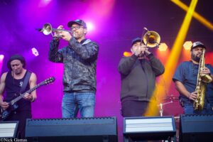 Band performance on stage: three musicians—two trumpeters and a guitarist—under vibrant purple and yellow lights, with a saxophonist on the right side, all playing.