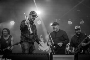 Black-and-white photo of a brass band performing on stage, center man in cap playing trumpet with other musicians nearby under bright stage lights