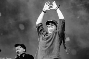 Concertgoer in a plaid shirt and sunglasses raises arms to form a heart shape above his head at a performance.