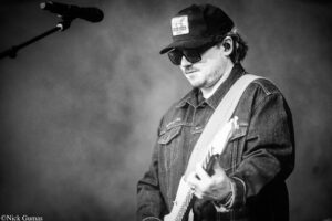Black-and-white photo of a guitarist wearing a denim jacket and cap, playing a white electric guitar on stage.