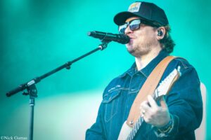 Guitarist and singer wearing a denim shirt, cap, and sunglasses, performing into a microphone on stage with a teal background.