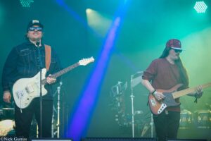 Two musicians on an outdoor stage: left guitarist in denim jacket and cap with a white electric guitar, right bassist in a maroon sweater and cap under blue-green lights.