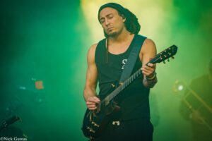 Guitarist with dreadlocks wearing a black sleeveless shirt, playing a black electric guitar on a green-lit stage during a live performance.