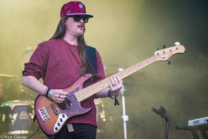 Bass guitarist in a maroon sweater and floral red hat plays on stage with a sunburst bass guitar--live performance.