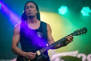 Guitarist with a beanie and sleeveless shirt playing a black electric guitar on a colorful stage, eyes closed.