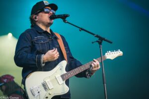 Musician in a denim jacket and cap sings into a microphone while playing a white electric guitar on a teal-lit stage.