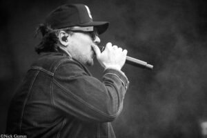 Profile view of a male singer wearing a backwards cap and denim jacket, singing into a handheld microphone on stage (black-and-white).