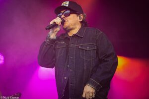 Male singer on stage wearing a denim jacket, cap, and sunglasses, singing into a handheld microphone under pink-purple lights.