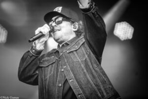 Male singer wearing a cap and sunglasses, microphone in hand, raising his fist on stage during a concert (black and white).