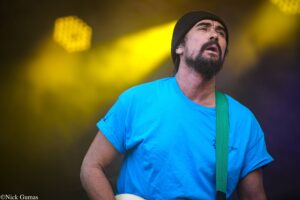 Bearded man in a blue shirt and black beanie on stage, wearing a green guitar strap under yellow stage lights.