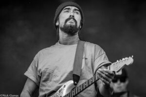 Bearded guitarist wearing a beanie, playing an electric guitar on stage, looking upward.
