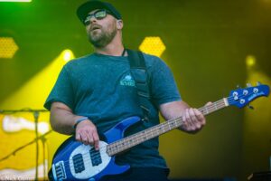 Musician playing a blue bass guitar on stage, wearing a cap and sunglasses amid green-yellow stage lights