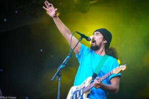 Male guitarist and singer wearing a blue T-shirt and black beanie performs on stage, raising his left hand as he sings into a microphone with a yellow spotlight behind him.