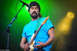 Male guitarist in a blue t-shirt and black beanie playing an electric guitar on a green-lit stage with a microphone nearby.