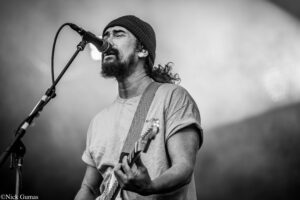 Male musician wearing a beanie sings into a microphone while playing an electric guitar on stage (black and white).