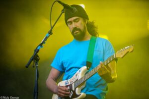 Musician in a blue T-shirt and beanie plays a cream Fender Strat on stage with yellow lighting on him.