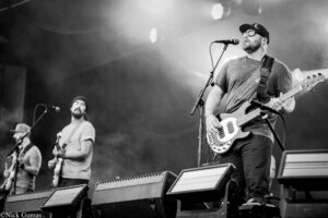 Black-and-white photo of a bass guitarist on stage, wearing a cap and sunglasses, singing into a microphone while other band members appear in the background.