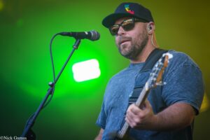 Male musician wearing a cap and sunglasses sings into a microphone on stage while playing a guitar.