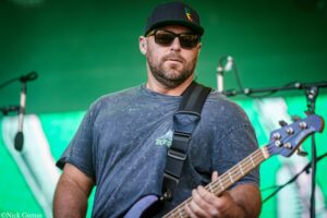 Male guitarist wearing a black cap and sunglasses, playing a bass on a green stage backdrop.