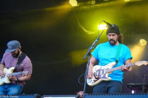 Two guitarists performing on stage under yellow lights; one in a blue shirt with a green strap, the other in a purple shirt and gray cap.