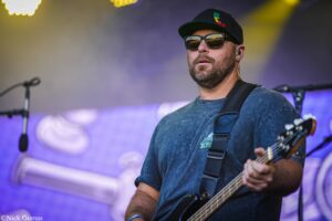 Male bass guitarist on stage, wearing sunglasses and a black cap, playing his instrument with a microphone stand blurred in the foreground and colorful stage lights behind him.