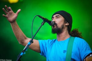 Male singer wearing a black beanie and blue shirt, singing into a microphone with one hand raised on a green stage background.