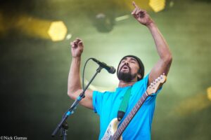 Male musician on stage singing into a microphone while playing an electric guitar, arms raised in the air.