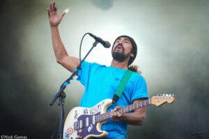 Male guitarist singing into a microphone on stage, wearing a bright blue shirt and black beanie, with a sticker-covered electric guitar in view.