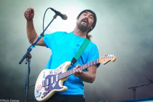 Guitarist in a bright blue shirt performing on stage, singing into a microphone while playing a sticker-covered cream Fender Stratocaster at a live concert.