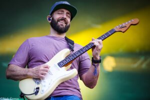Bearded guitarist in a purple shirt and cap smiling while playing a white electric guitar on stage with yellow background light.