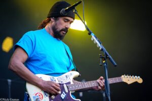 Guitarist in a blue shirt and black knit hat playing a white Fender Strat on stage with a microphone in front of him.