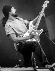 Black-and-white photo of a bearded guitarist in a knit cap, intensely playing a decorated Fender Stratocaster on stage.