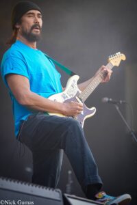 Guitarist on stage wearing a blue shirt and black beanie, playing a white sticker-covered electric guitar with a green strap.