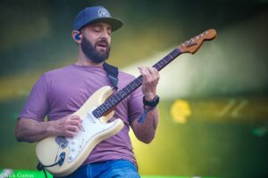 Male guitarist in a blue cap and purple shirt plays a cream Fender Stratocaster outdoors, eyes closed as he performs.