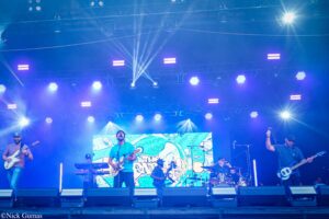 Live band performing on a blue-lit stage with musicians playing guitars and drums, behind a colorful screen backdrop.
