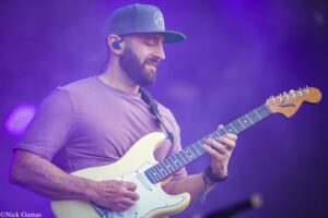 Guitarist wearing a cap and purple shirt plays a white electric guitar on stage with purple lighting in the background