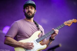 Bearded guitarist wearing a cap plays a cream Fender Stratocaster on stage with purple lighting in the background.